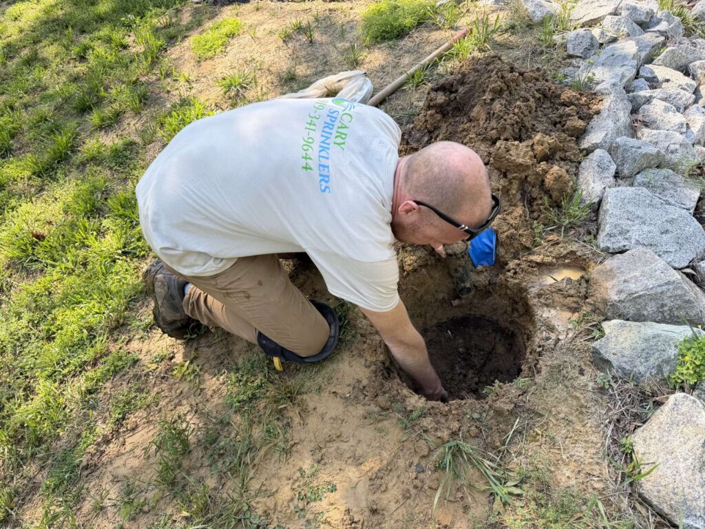 Technician repairing an irrigation line on a sloped, rock-bordered yard in the Cary, NC area