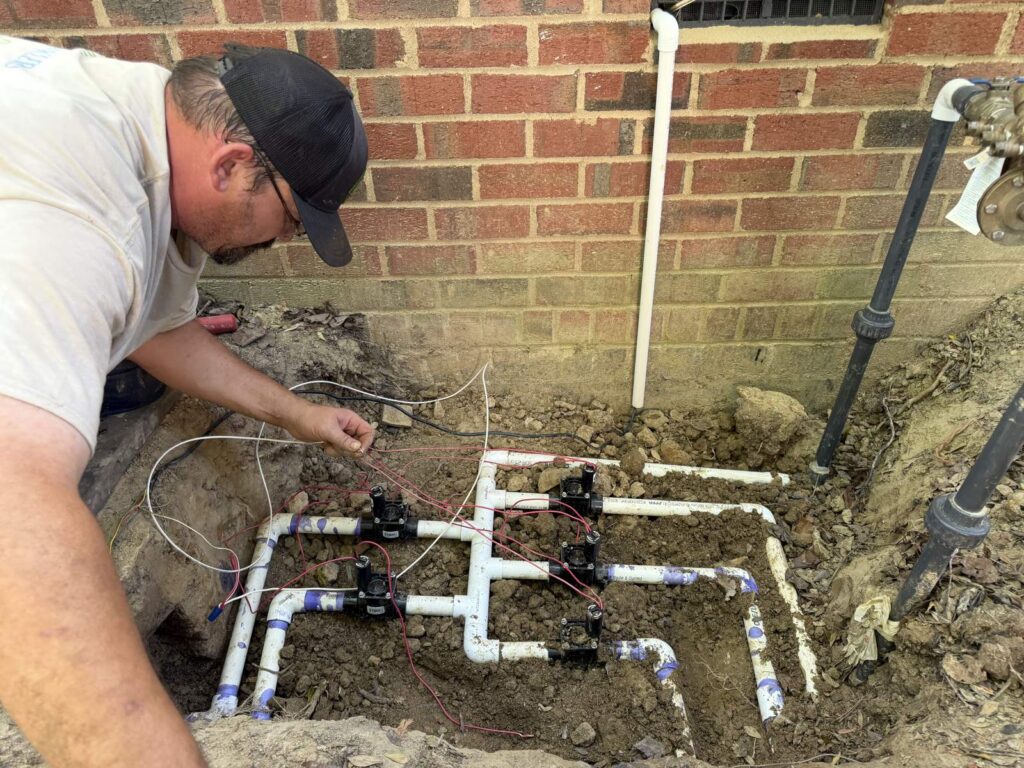Technician rebuilding a sprinkler valve manifold against a brick wall in Cary, NC