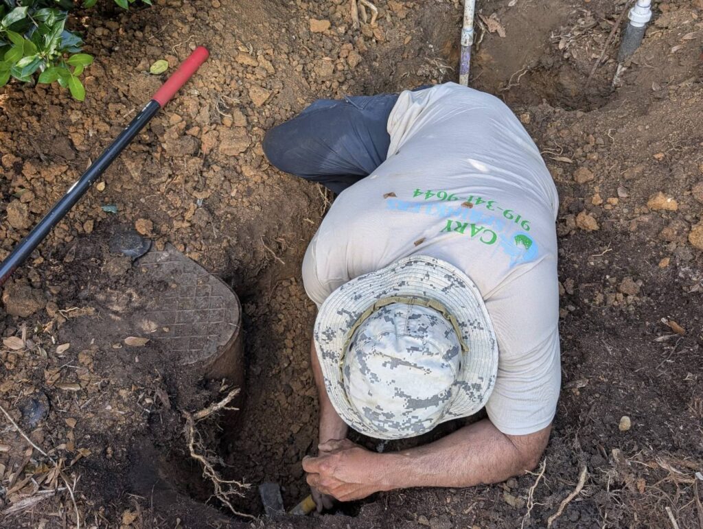 Technician reaching into an excavated valve box to repair an underground irrigation leak in Cary, NC